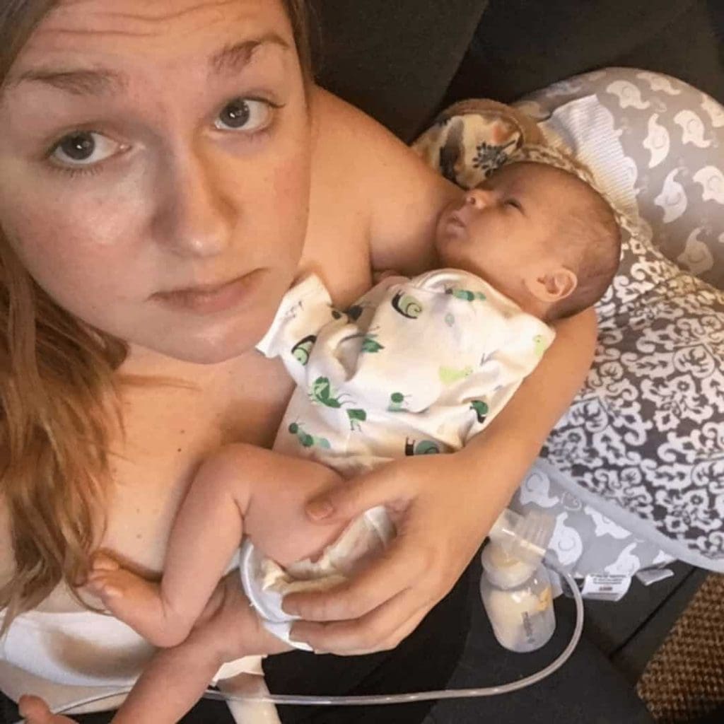 woman-looking-up-at-camera-holding-baby-on-arm-with-breast-pump-bottles-under-baby