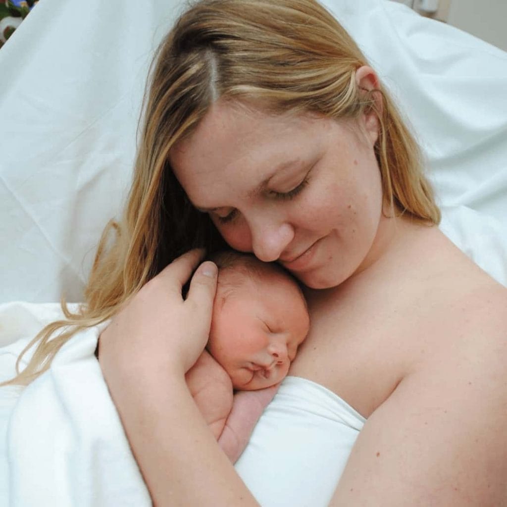 new-mom-wrapped-in-white-sheet-holding-newborn-baby-against-chest-with-smile