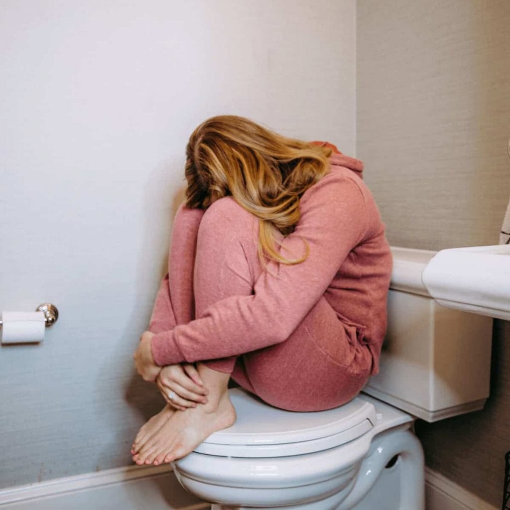 woman sitting on closed toilet with her arms wrapped around her legs and her head buried