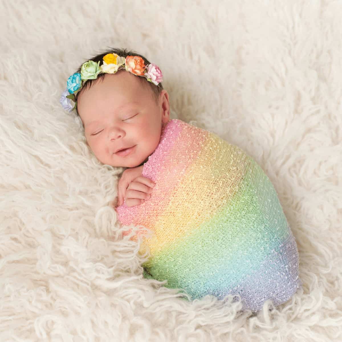 newborn-baby-with-dark-hair-and-rainbow-colored-flower-headband-lying-on-fuzzy-blanket-swaddled-in-rainbow-blanket