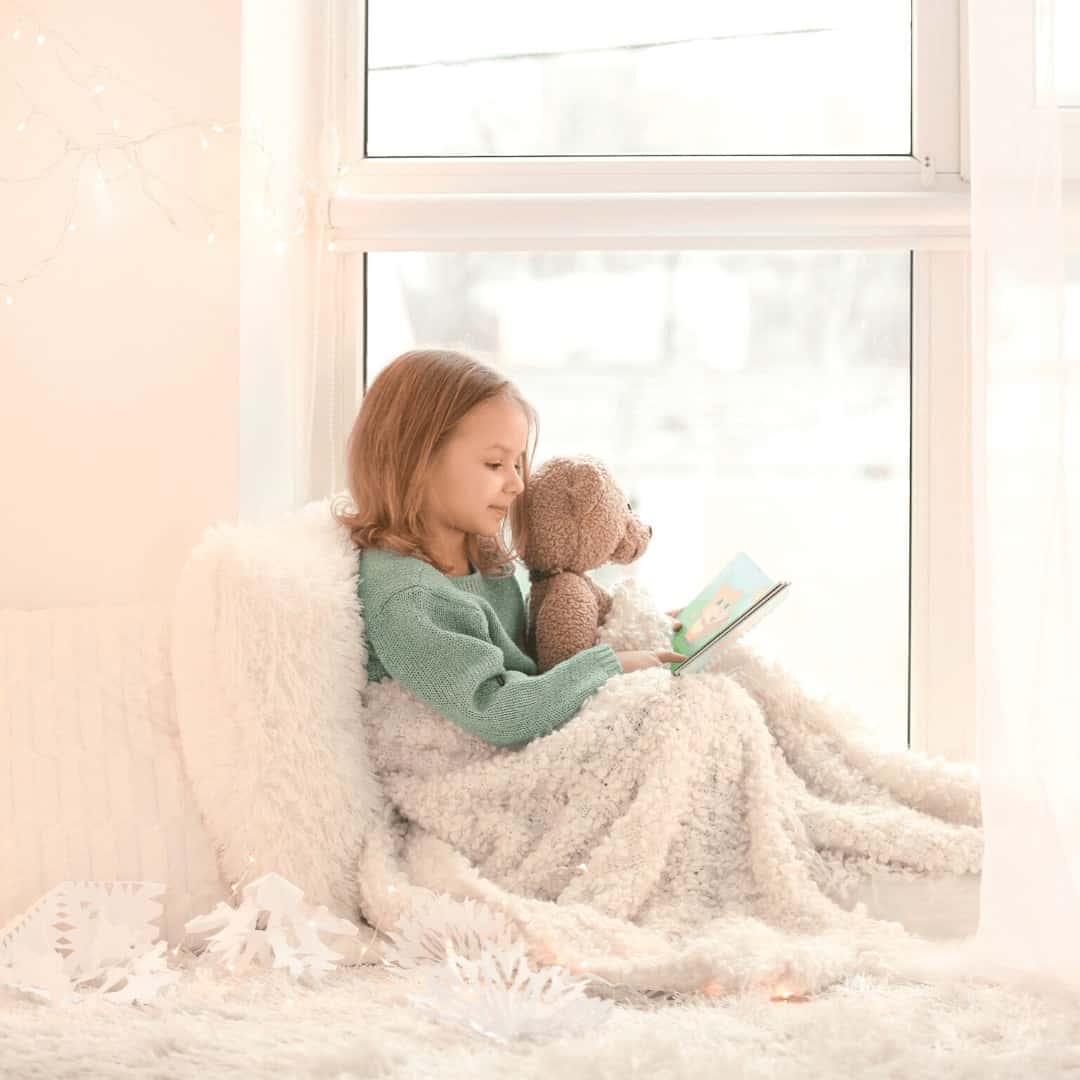 little-girl-sitting-in-window-seat-snuggled-in-winter-blankets-holding-teddy-bear-reading-a-book