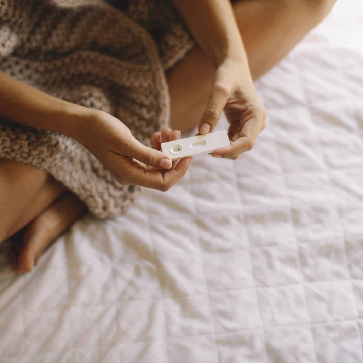 woman's hands holding negative pregnancy test sitting under a blanket on a bed