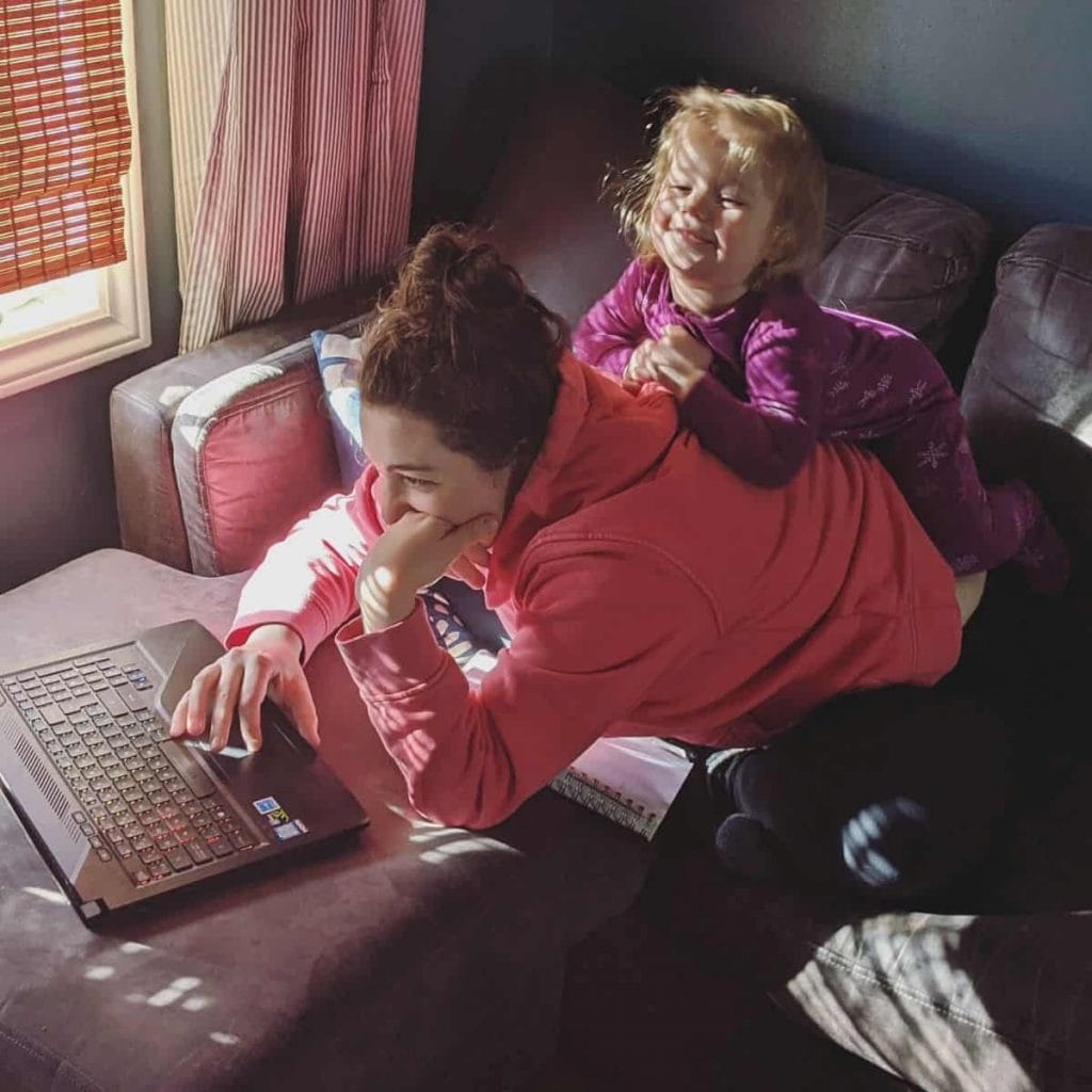 mom leans forward and works on computer while her child plays on her back