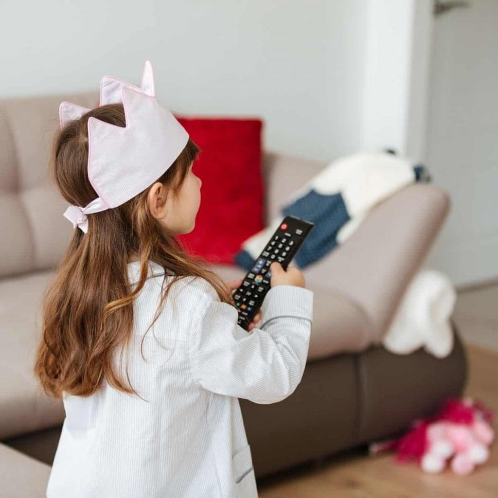 Back view of little girl using remote to turn on the TV in living room at home