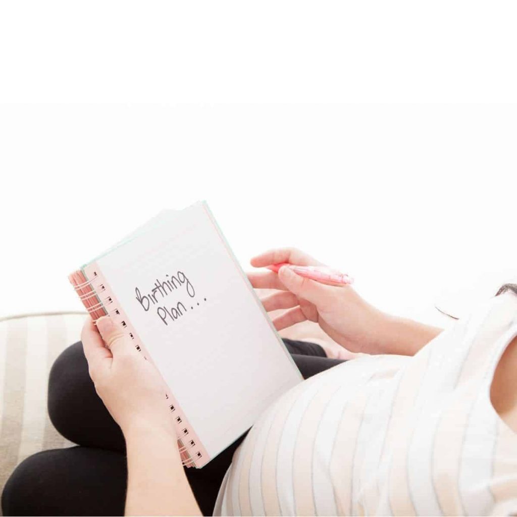 woman with pregnant belly sitting on couch holding notebook with the words birthing plan dot dot dot on an otherwise blank page