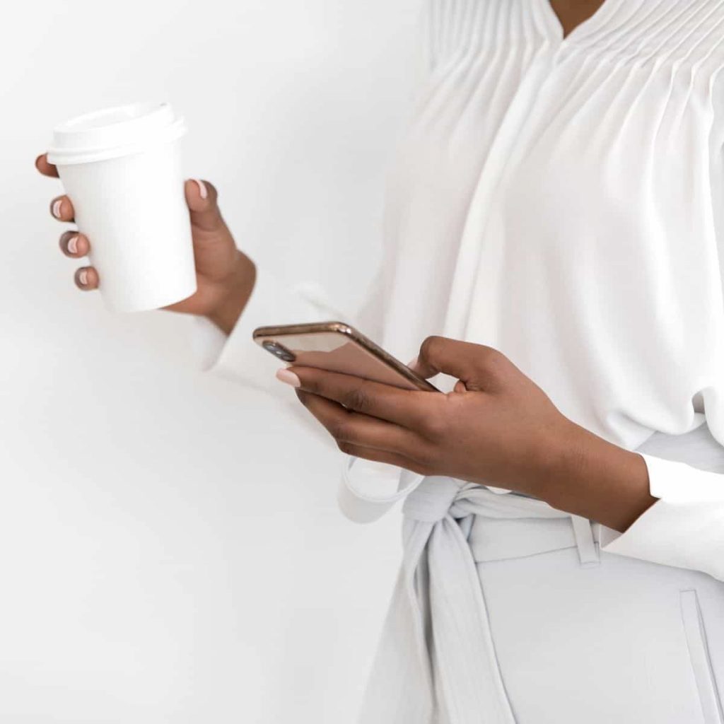 women wearing white blouse is looking down at her iPhone in one hand and holding a coffee cup in her other hand