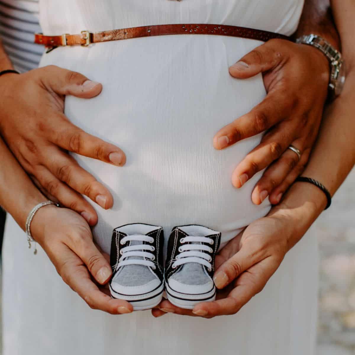 photo showing mother and father's hands; the father is holding the pregnant belly and the mother is holding baby shoes in front of her stomach