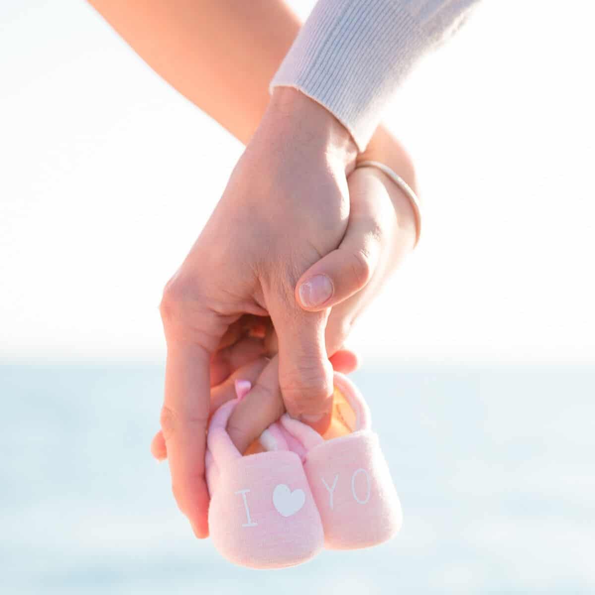 mother and father holding hands while clutching pink baby shoes