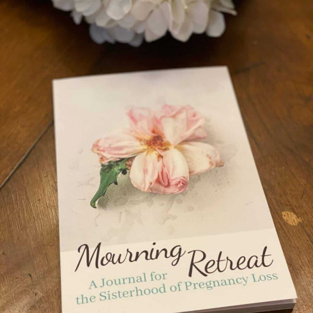 beautiful miscarriage journal called Mourning Retreat a journal for the sisterhood of pregnancy loss lying on a wooden surface with a beautiful flower on the cover