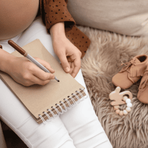 A pregnant individual sits with a notepad on their lap, poised to write. The person is wearing a brown polka-dot shirt and white pants, and they are partially shown, with the image cropped to exclude their head. To their right, on a fluffy beige rug, there are baby accessories: a pair of tiny brown lace-up shoes and a wooden toy. The scene suggests a peaceful moment of contemplation, possibly related to choosing a name for the expected baby, as indicated by the items and the notepad.