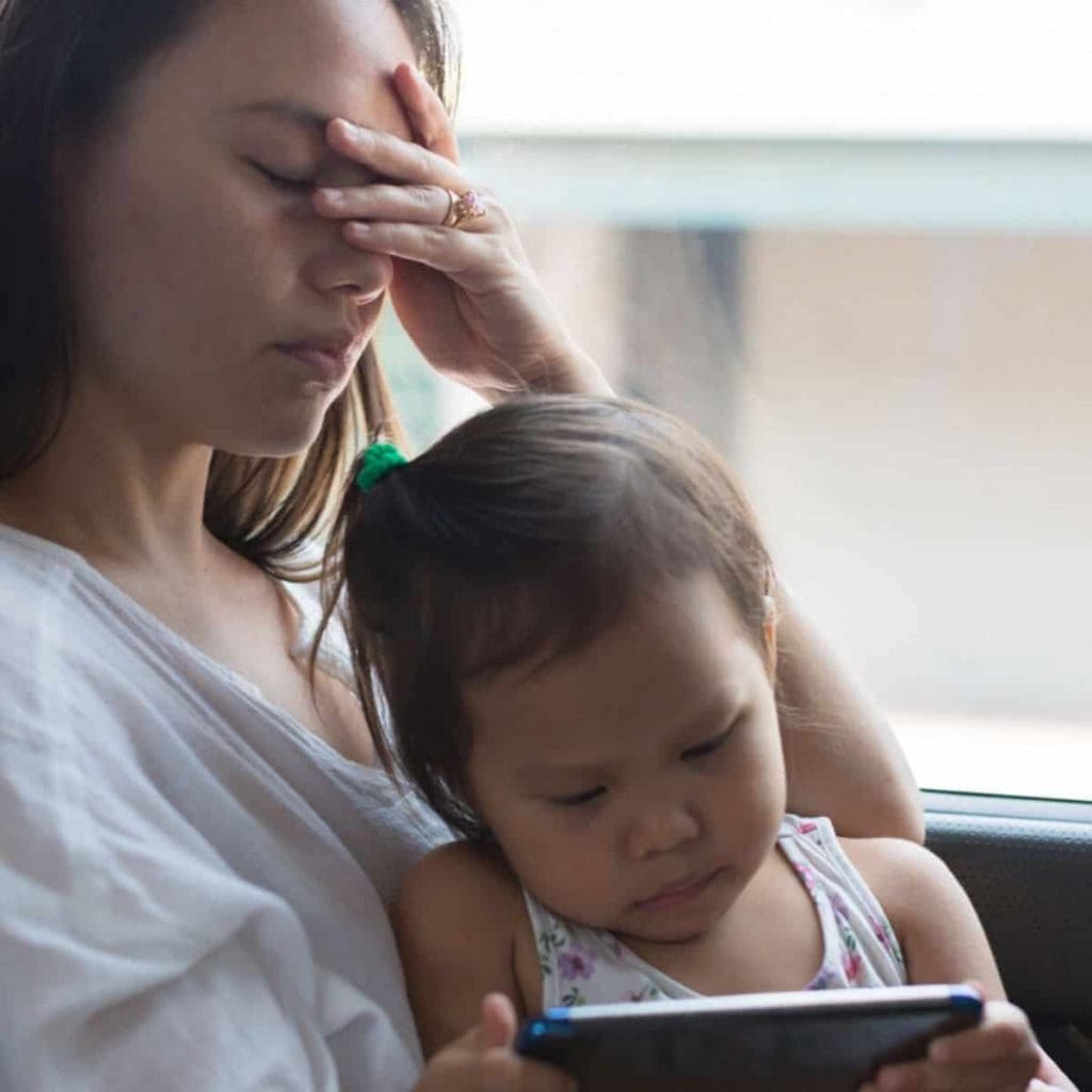 overwhelmed mom holds her face in her hands while her toddler daughter plays on a tablet