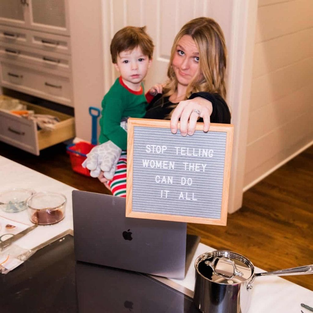 Mom holding toddler and a letterboard that reads "Stop Telling Women They Can Do It All"