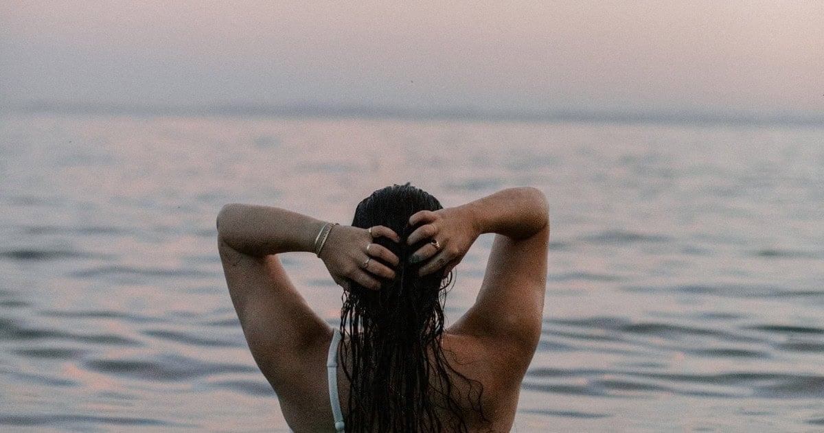 A woman stands waist-deep in calm ocean water at dusk, seen from behind, wringing out her wet hair with both hands as a soft pink-and-lavender sunset stretches across the horizon. A quiet, unposed moment of a woman alone with her body and the water, ahead of a roundup of the best postpartum swimsuits.