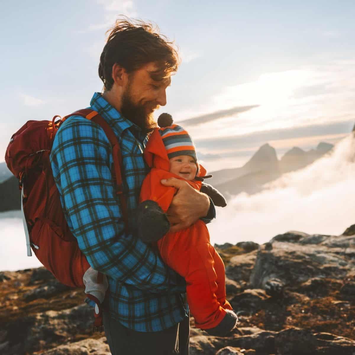 dad wearing a diaper backpack and holding baby while vacationing in the mountains