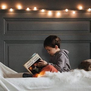 Toddler is reading a book while in bed with nightlights spread out on headboard