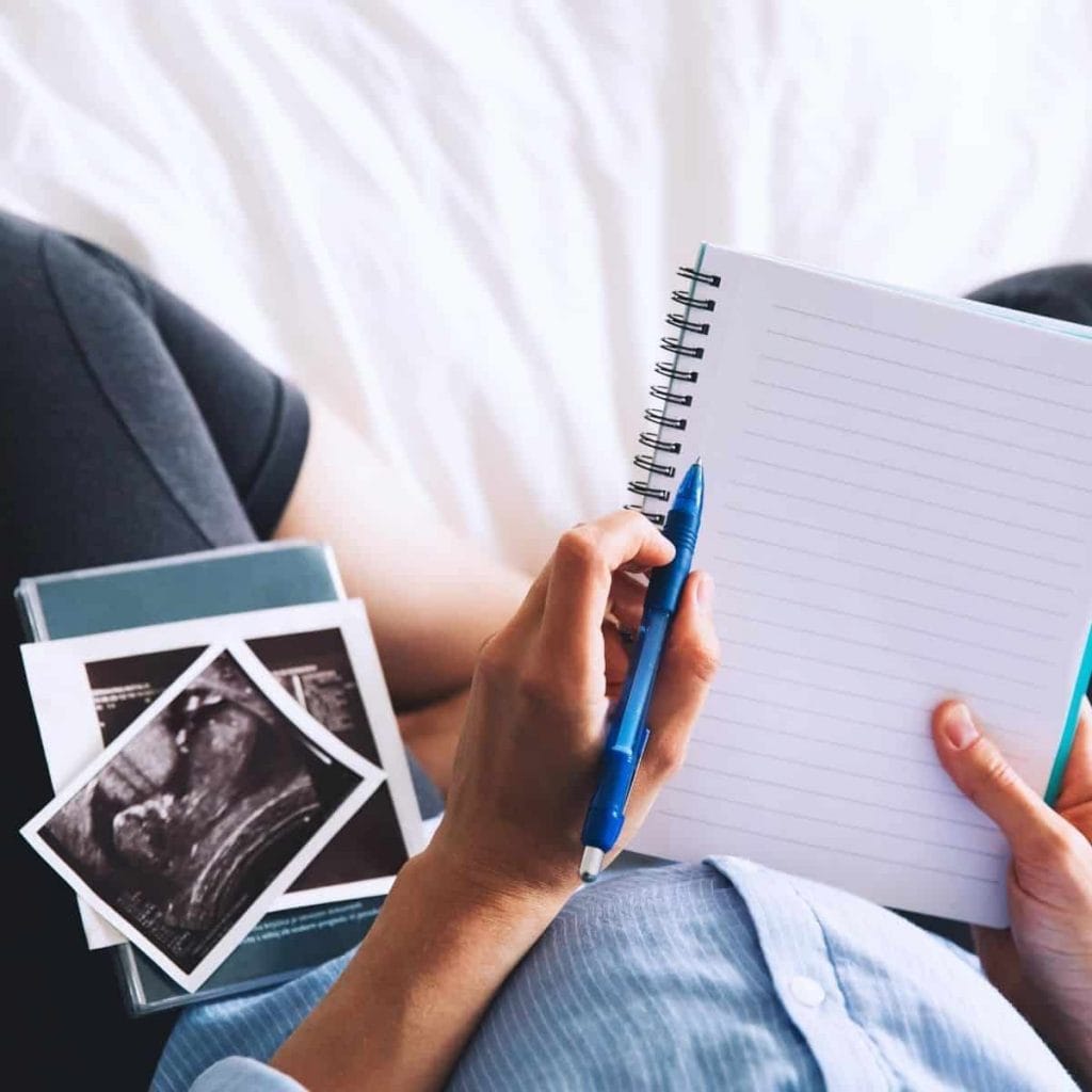 pregnant woman writing on notepad with a sonogram picture in laying on her lap