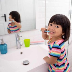 young toddler brushes her teeth in front of bathroom sink with her reflection in mirror