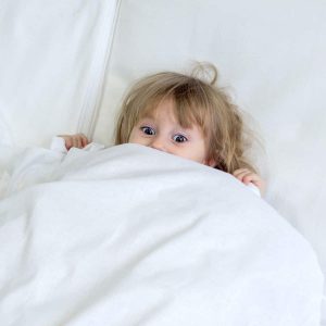 little girl hiding underneath white bedsheets with her eyes and hands showing