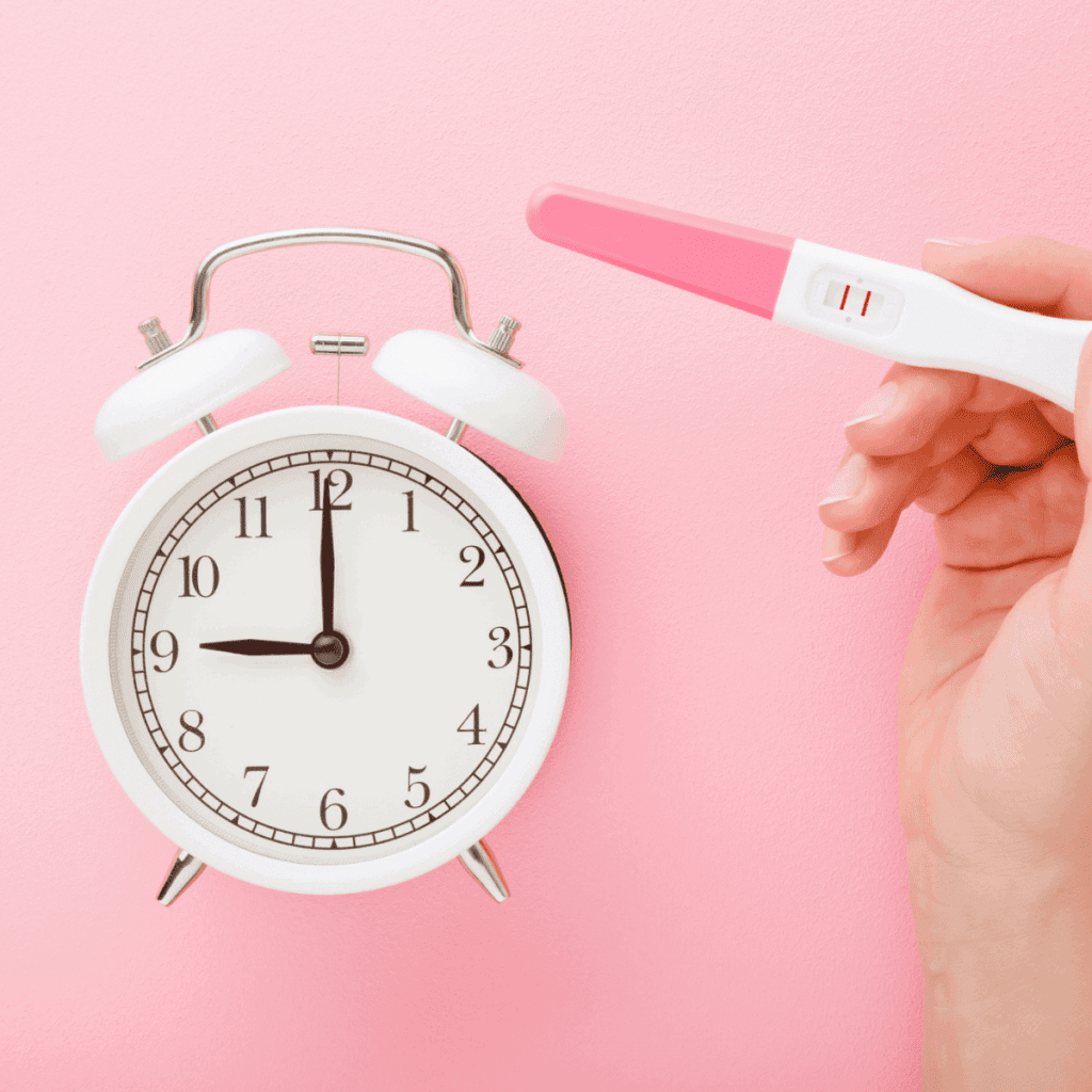 A white clock on a pink background with a woman's hand holding a positive pregnancy test next to the clock