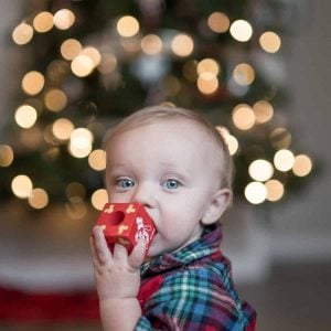 A baby with bright blue eyes chews on a Christmas ornament in front of a blurred Christmas tree with lights.