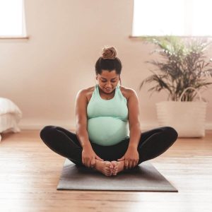 Pregnant woman sitting on yoga mat with feet touching in a light blue tank top and black leggings.