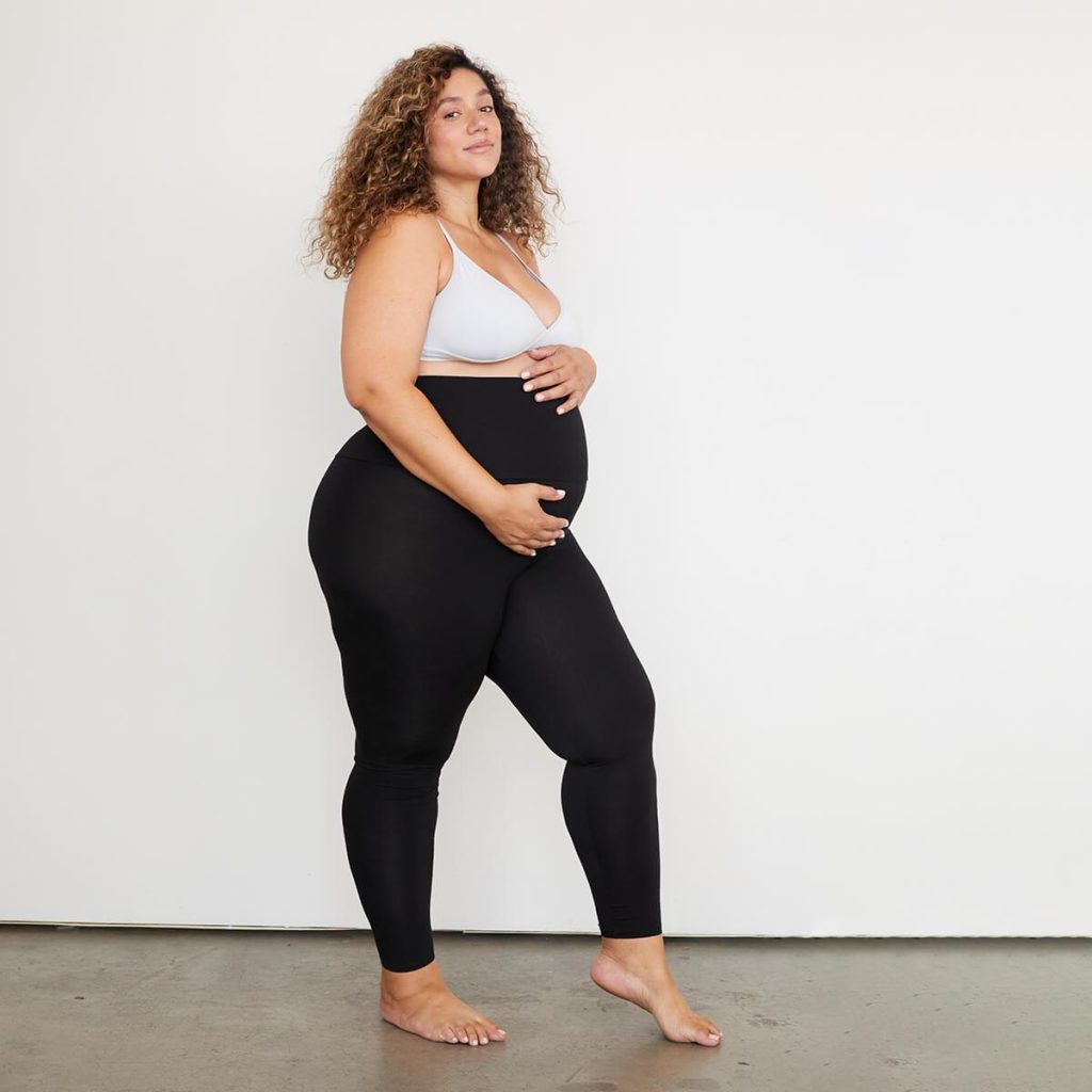 Woman with long dark curly hair stands in front of a white wall wearing black leggings with her arms showing off her pregnant stomach