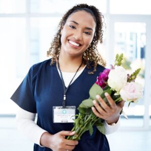 An African American woman with brown curly hair is wearing a nurse's uniform and smiling while holding a bouquet of pink and white flowers