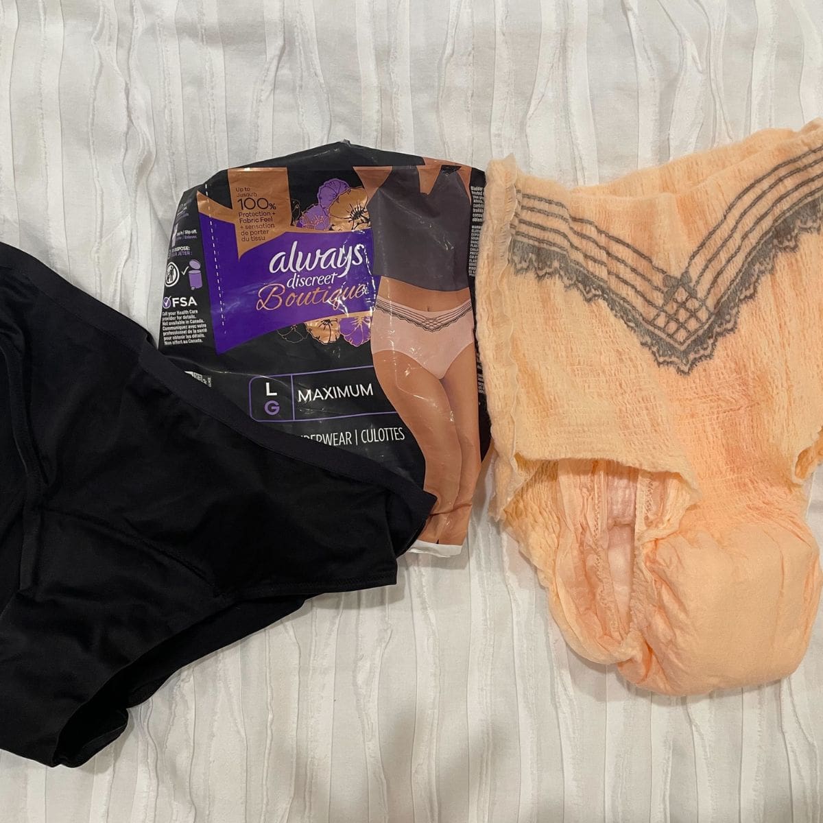 A pack of always discreet boutique women's adult diapers lie on a bed with a diaper open to its right and a pair of black Thinx period panties to its lef.