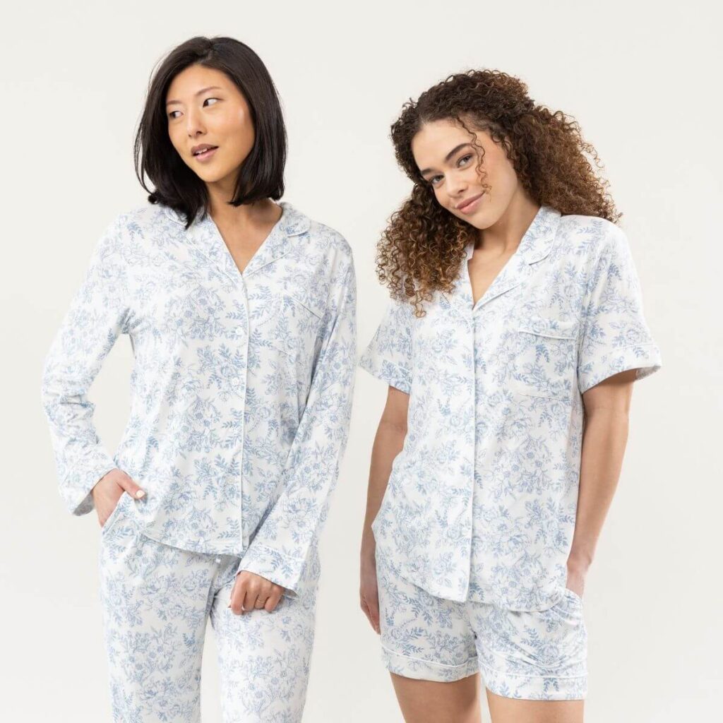 2 women stand together in front of a white background. The woman on the left is taller, a beautiful woman of Asian descent with shoulder-length hair, wearing a long-sleeved button up bamboo pajama set. The pajamas are white with a soft blue floral pattern. The woman on the left is slightly shorter, an African American woman with light skin and reddish-brown hair, wearing the same pajama print, but she is in a short-sleeved pajama set.
