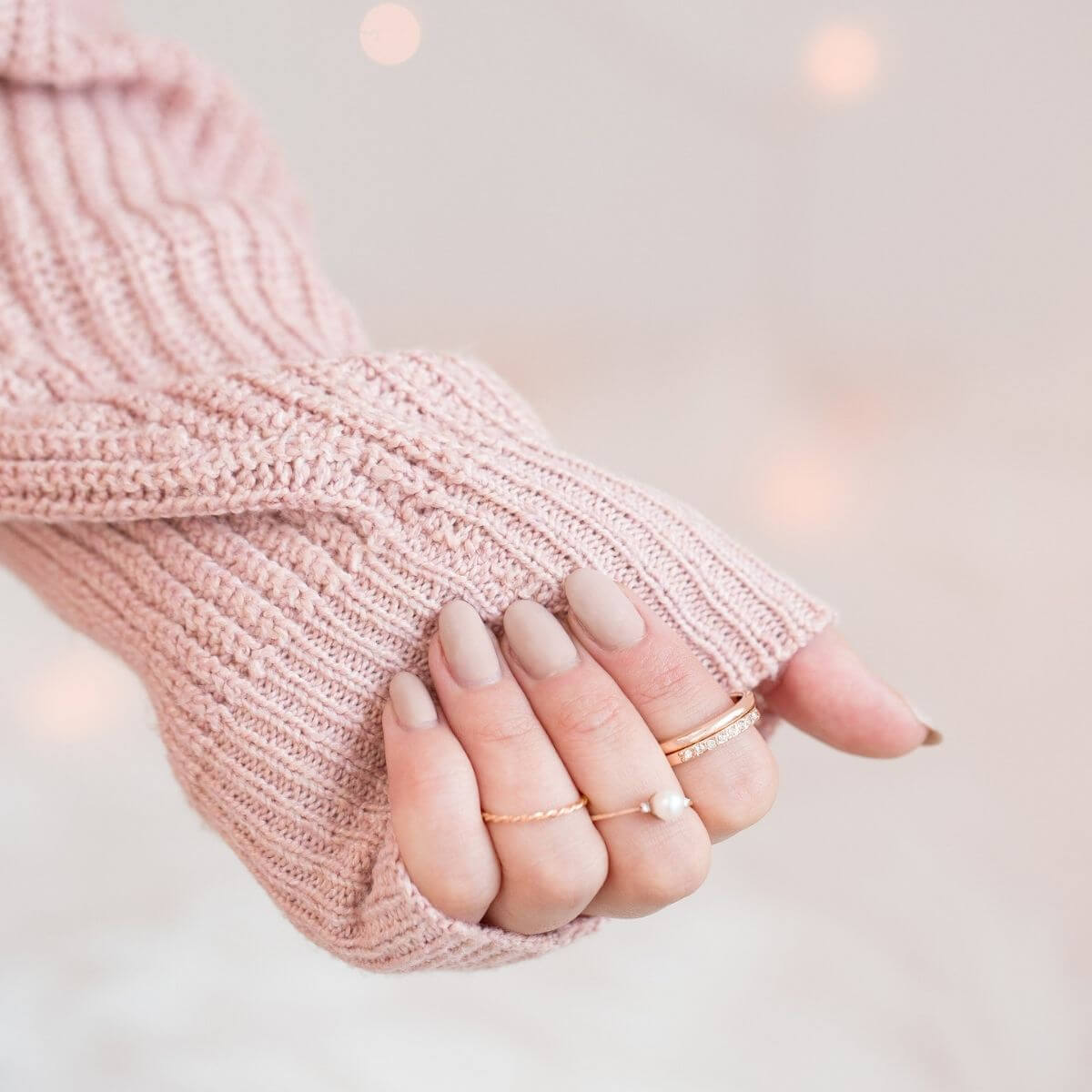 A close-up photo of a person's hands partially covered by a pink, knitted sleeve. The visible hand has neatly manicured nails painted in a soft, matte nude polish. On the ring finger, there's a delicate, simple gold band, and on the middle finger, there's a thin gold ring with a small pearl. The overall feel of the image is cozy and gentle, with a bokeh effect creating soft light circles in the background, suggesting a warm, tranquil environment.
