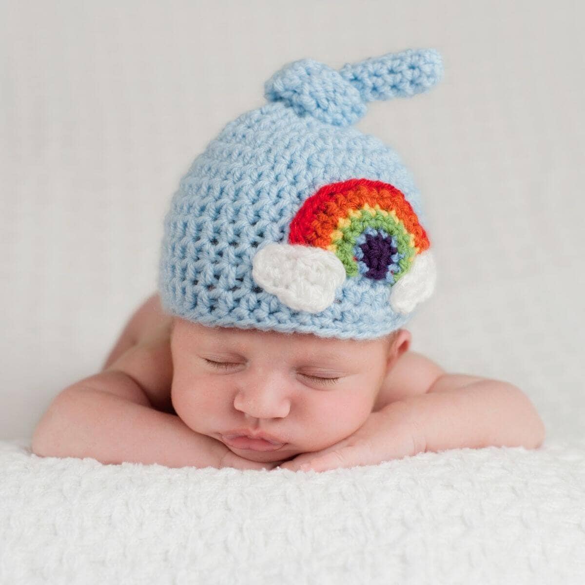 newborn baby lying with its chin on its hands wearing a blue knit hat with a rainbow on it