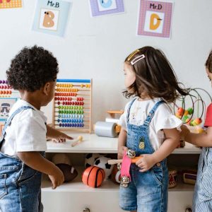 African American boy with a white shirt and overalls, a caucasian girl with white shirt and overalls, and a caucasian boy with a red shirt and overalls playing with toys with green, yellow, organge, and blue beads on them