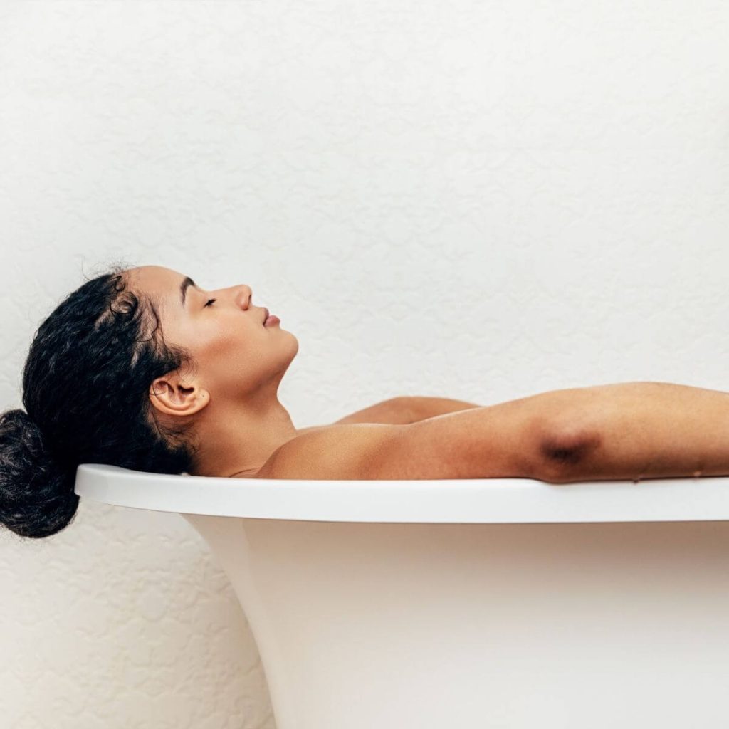 African American woman with hair in a bun laying back in a white tub with eyes closed and hands on the sides of the tub