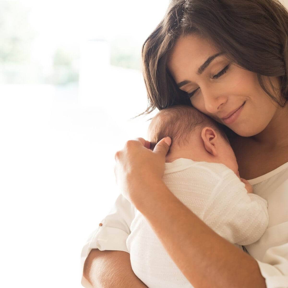 Caucasian woman with dark brown hair, eyes closed and smiling, in a white shirt snuggling a newborn baby in a white onesie to the side of her face