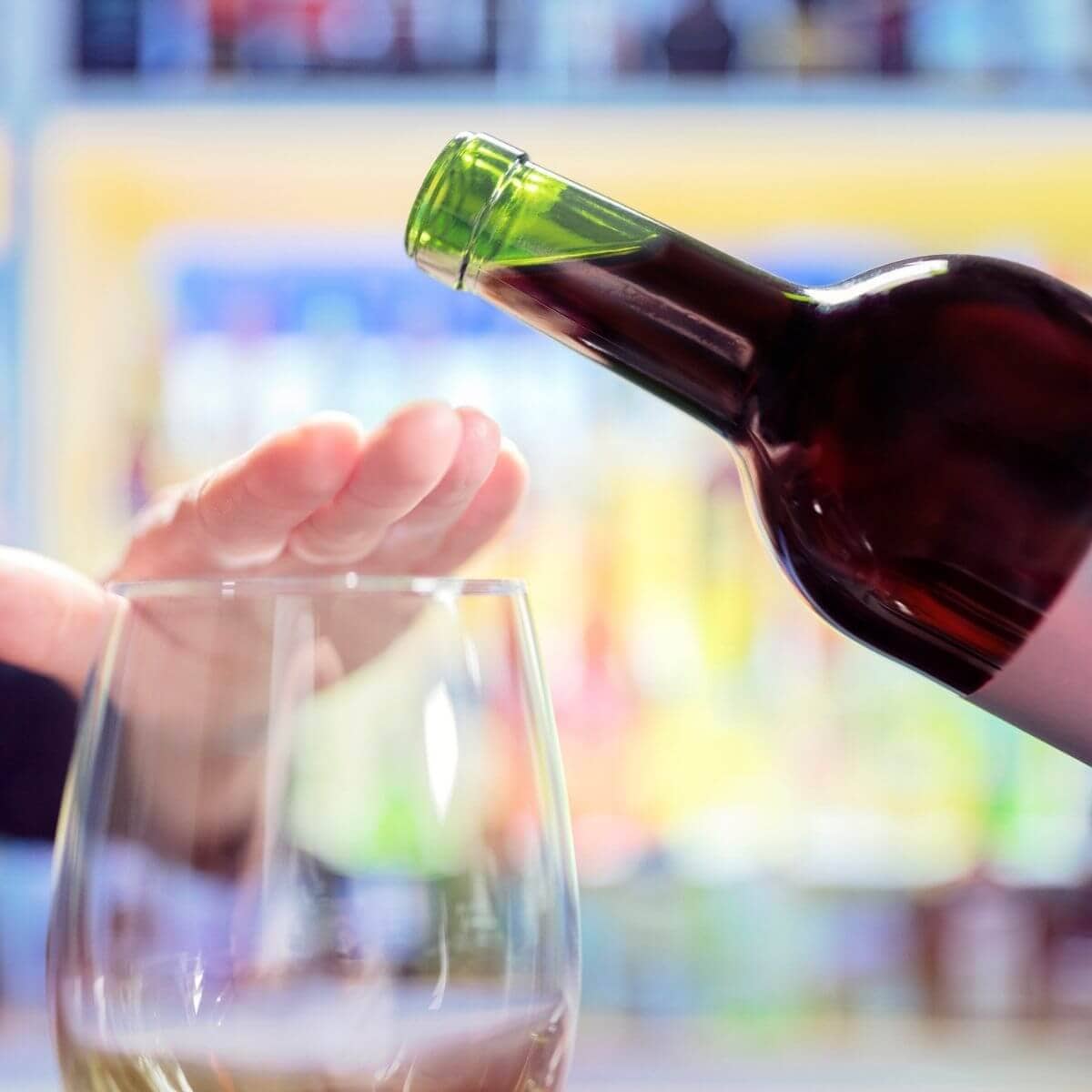 An empty stemless wine glass sitting on a table with a woman's hand over the top blocking wine from being poured in the glass while to the right of the glass is a green wine bottle with a dark red wine almost being poured over the hand that is covering the glass