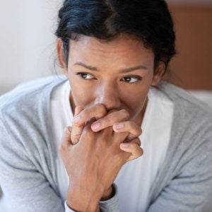 Woman with sad eyes sitting down wearing a white teeshirt and grey sweater with hands intertwined and in front of her mouth