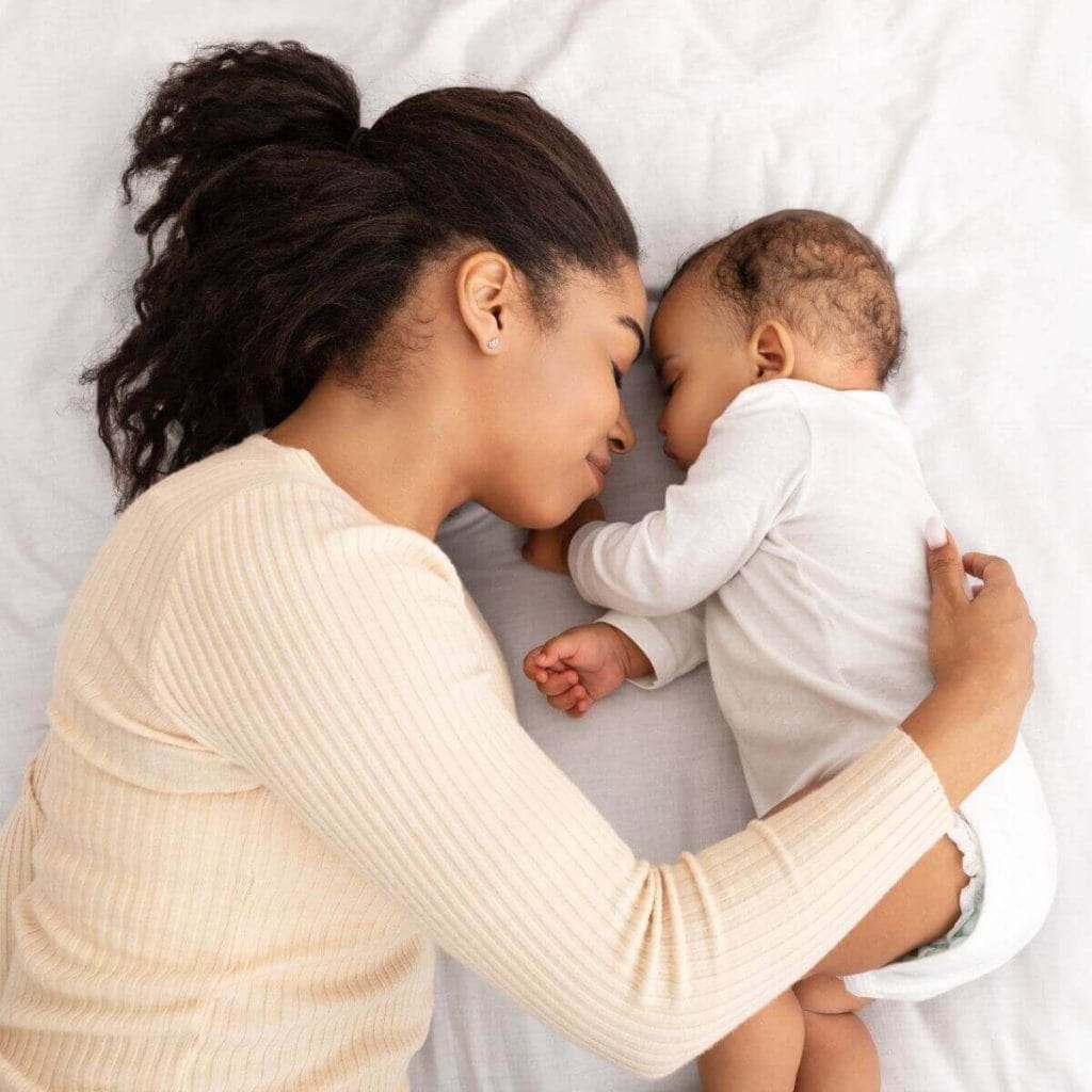 African American woman smiling with her eyes closed and wearing a light pink sweater laying on a bed with a white sheet over it with a newborn African American baby wearing a long sleeve white onesie sleeping and facing the woman