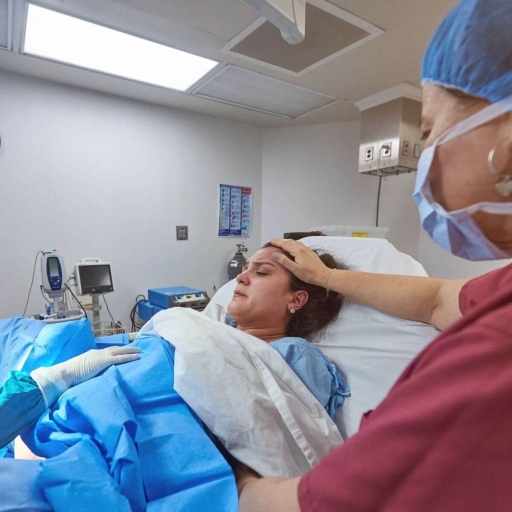 Woman laying on a hospital bed pushing out a baby and a nurse in rose colored scrubs with a blue head cover and blue and white mask on with her hand on the woman's head