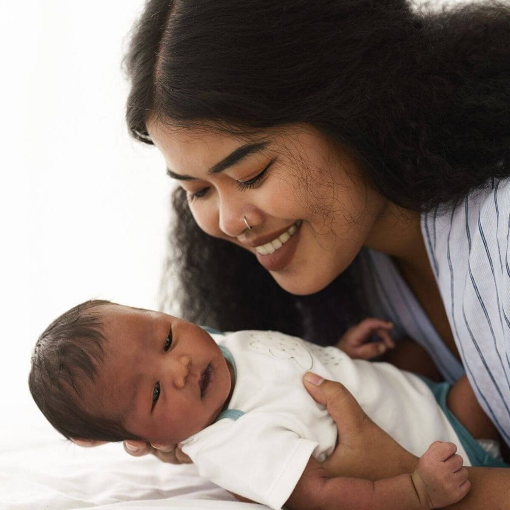 Woman with long black hair and a nose ring smiling down on a newborn baby in a white onesie with teal trim
