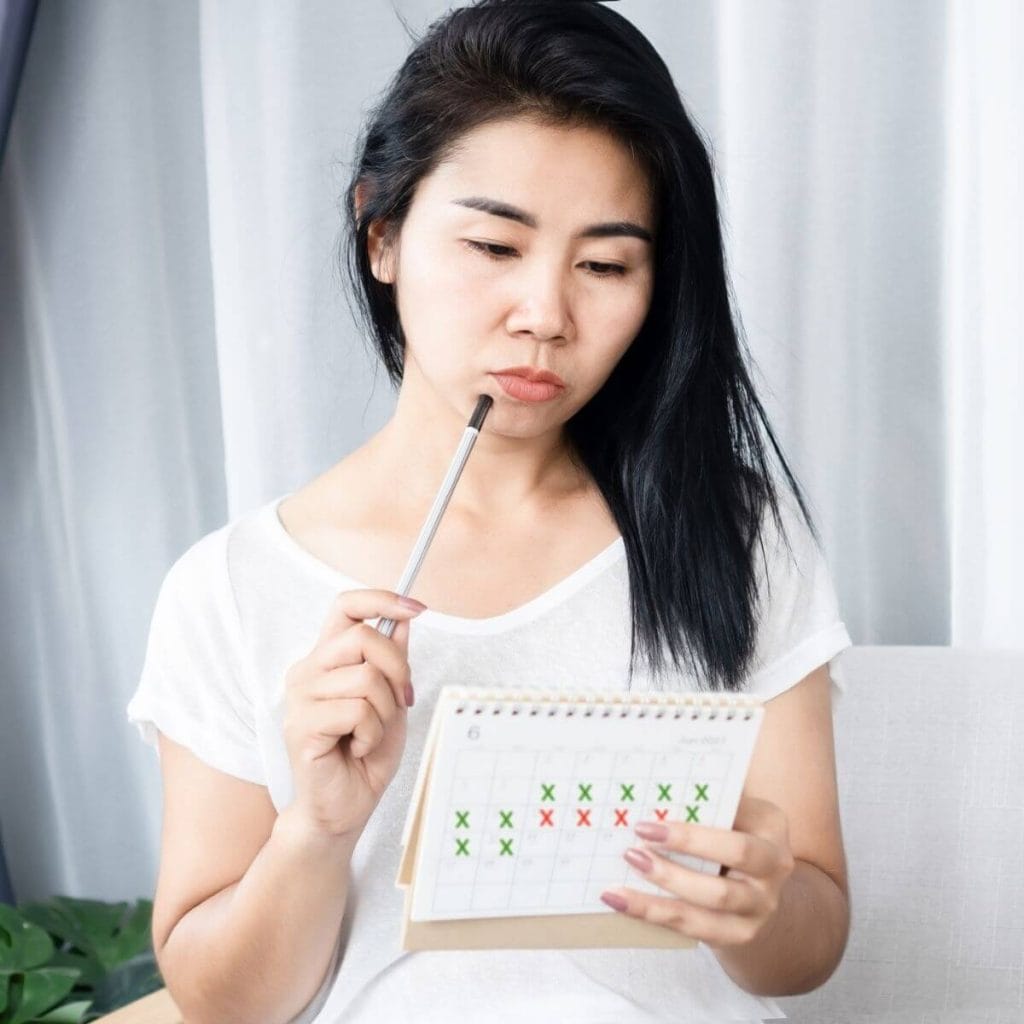 Asian American woman with long black hair sitting on a white couch with white curtains and a green plant behind her wearing a white t-shirt and looking thoughtfully at a calendar that has red and green x's on it