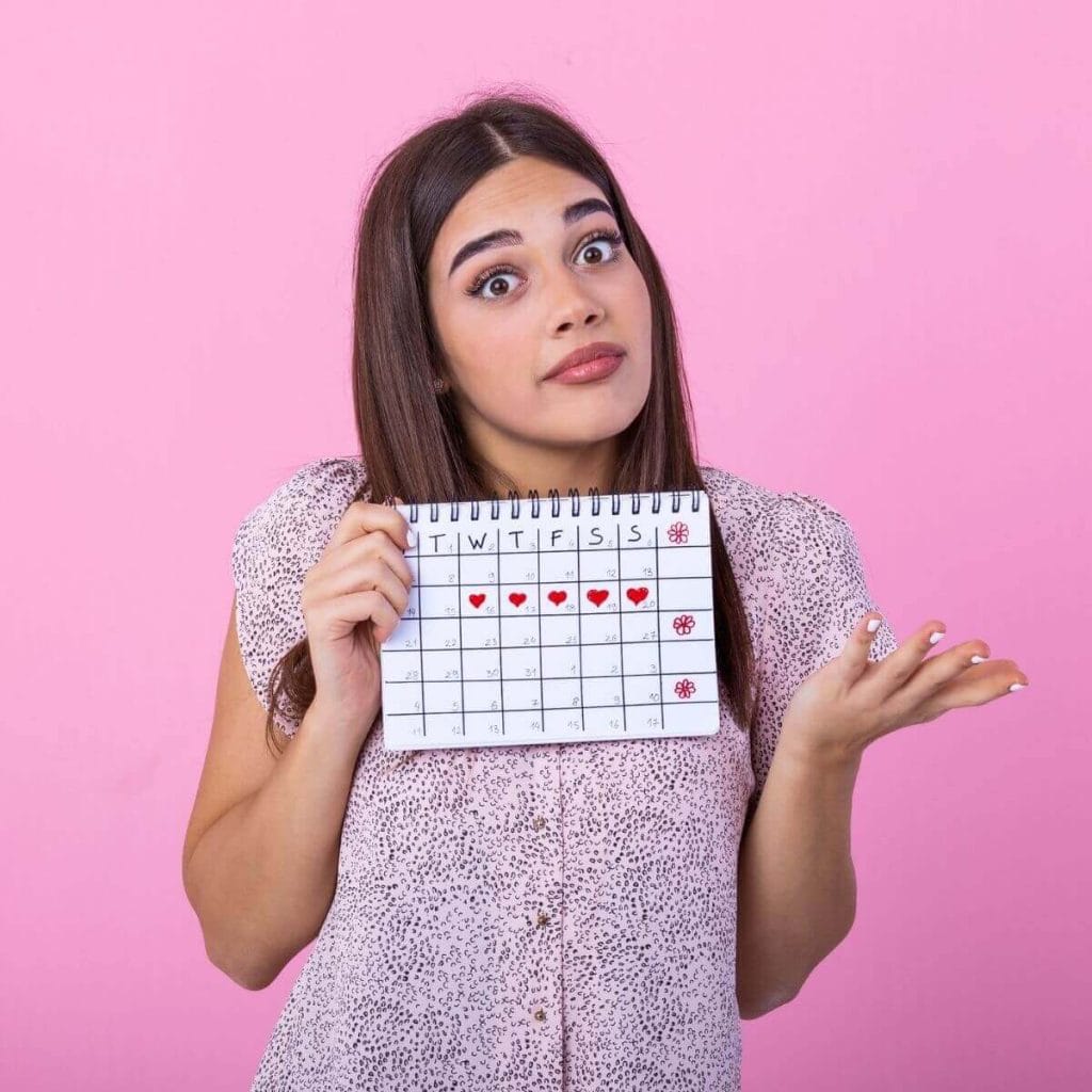 Caucasian woman with medium length dark brown hair in a white button up shirt with small dark purple dots all over it and white finger nails holding a calendar with 5 red hearts on it against a pink background with hands making the gesture "I don't know"