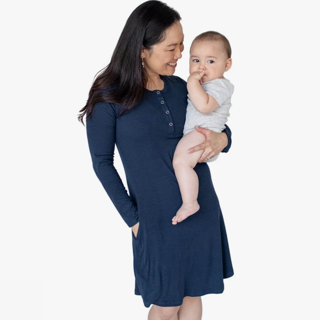 Woman wearing navy blue ribbed nightgown has one hand in pocket while holding her baby in the other