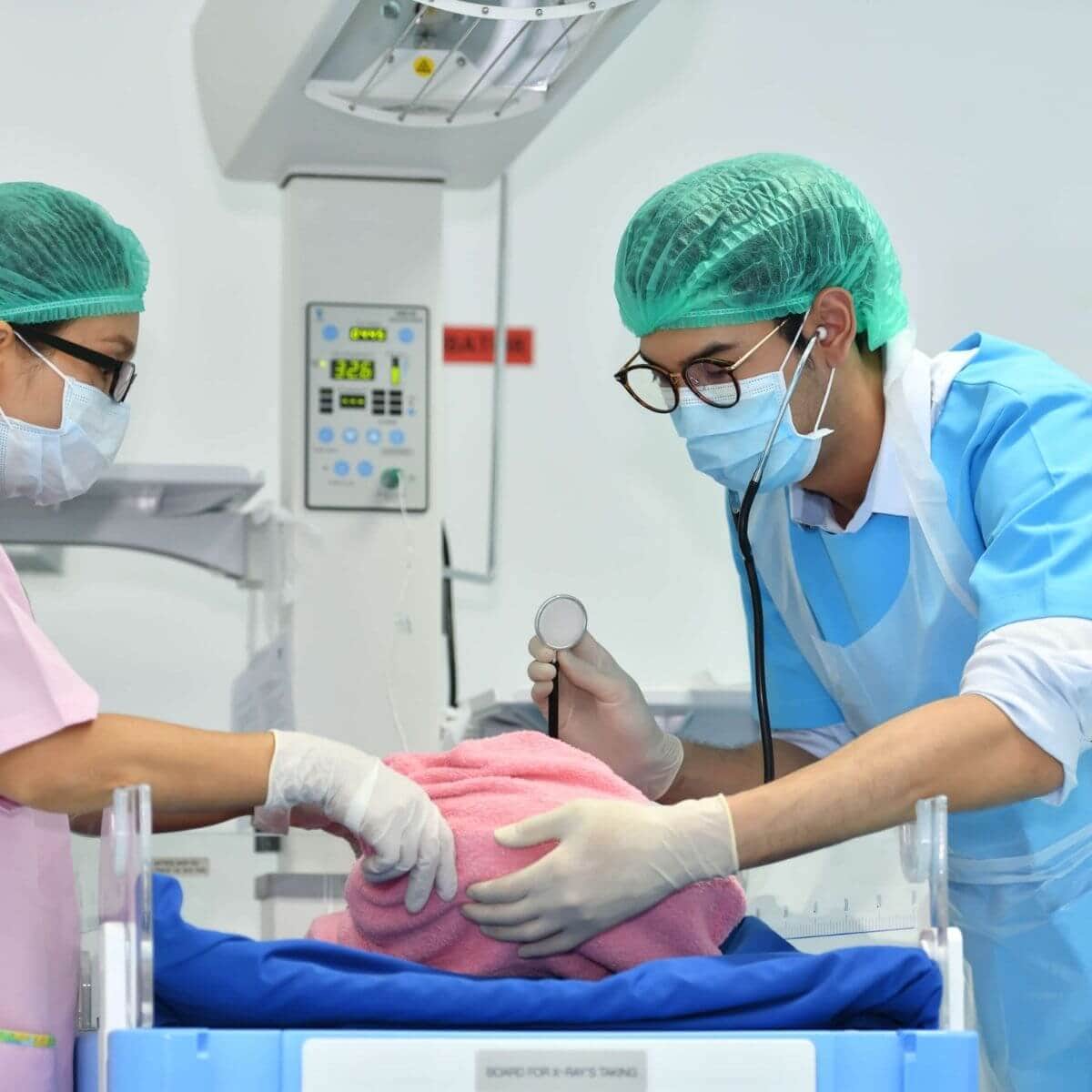 Newborn baby wrapped in a pink blanket on a hospital machine getting vitals checked by two Asian American nurses, one woman with light pink scrubs, a white mask and green hairnet and a male with light blue scrubs with a blue mask and green hairnet