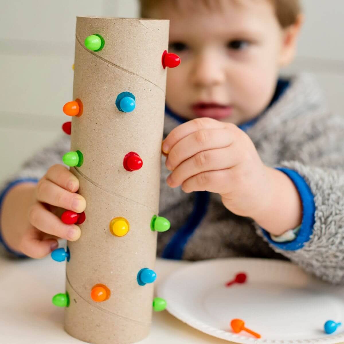 Close up shot on a little boy playing with a paper towel roll that has little holes cut it in, with different colored dots sticking out of each hole.