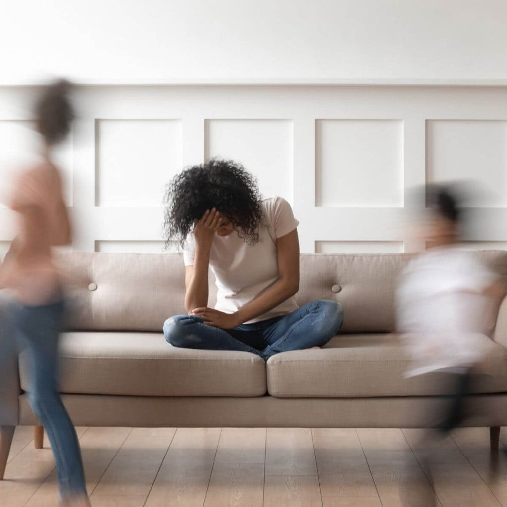 African American woman sitting on a light tan couch with hardwood floor and a white wall behind her head in hand white two kids run around in a blur