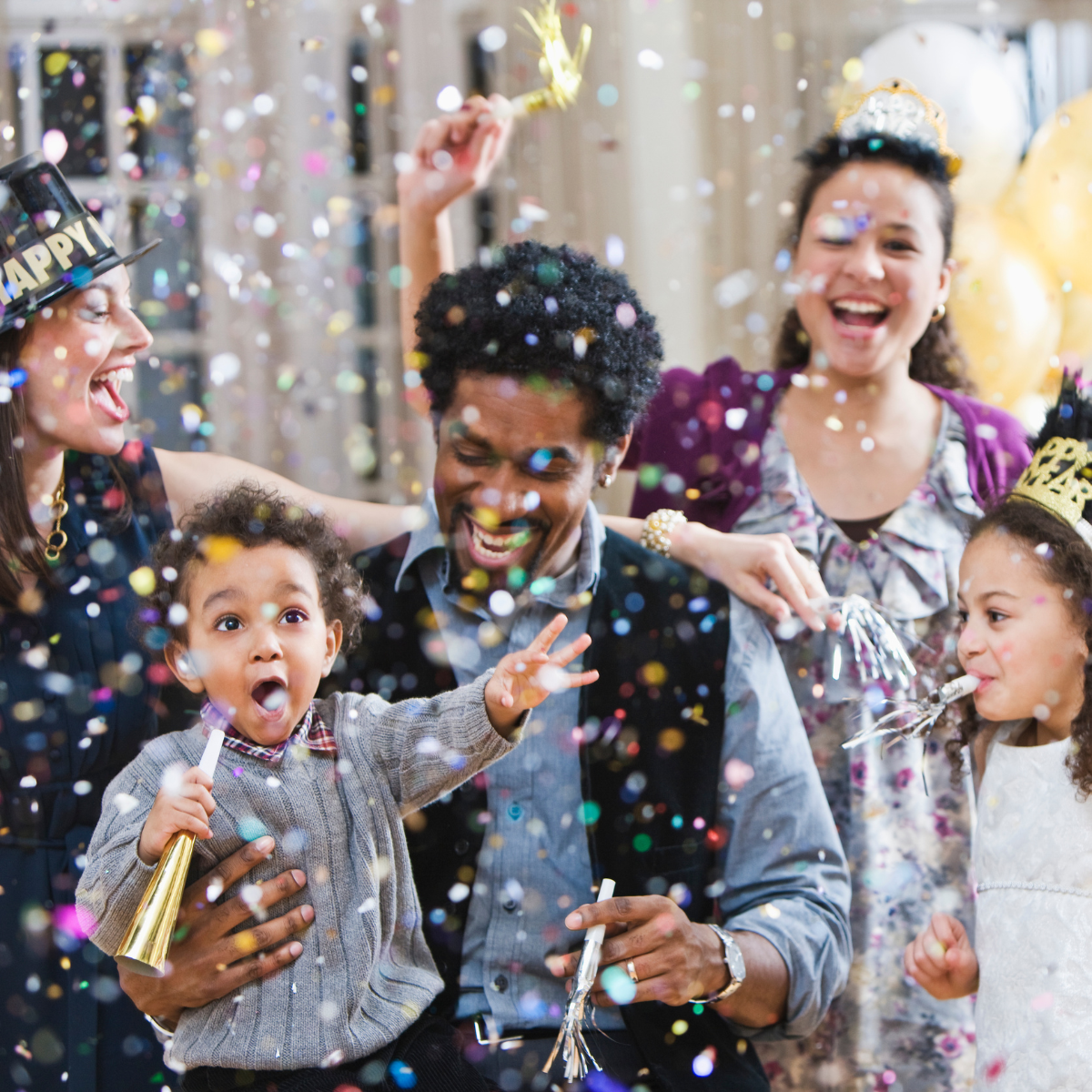 An exuberant family celebration with two adults and two children enjoying a New Year's party. The adults are wearing festive 'Happy New Year' hats, with one blowing on a party horn. A man and a young girl are playfully tossing confetti, while a toddler, held by the man, is wide-eyed with excitement, mouth open in awe. They are all surrounded by a shower of colorful confetti, creating a joyful and lively atmosphere. The backdrop suggests an indoor setting with balloons, adding to the festive environment.