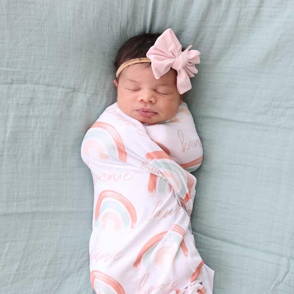 Baby lying on a soft blue blanket with a pink headband wrapped in a soft white swaddle blanket with bright pastel rainbows and the name Bonnie printed on the blanket
