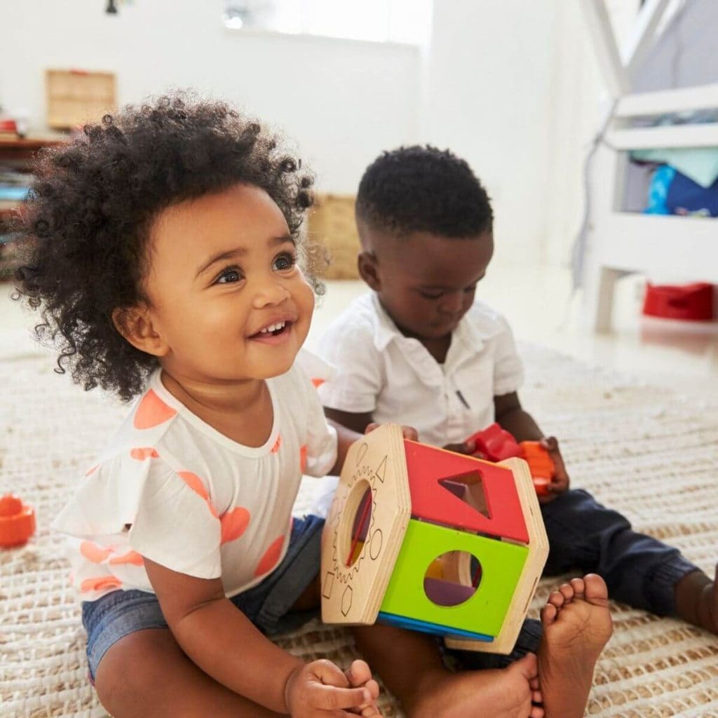 One year old African American boy sitting on a rug with a white shirt and jeans on holding one orange and one pink shape block with a 1 year old African American girl in a white and light orange poka dotted shirt and jean shorts smiles to someone off the camera while she is holding a box that the shapes fit into