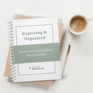 Spiral Bound Expecting & Organized Planner sitting on top of a light pink notebook which is laying on a table with a coffee mug full of coffee and a light pink pen sitting beside it