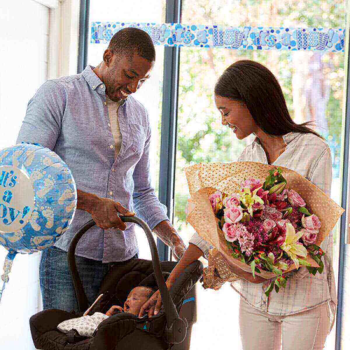 African American Man in jeans with a light blue button up shirt and an an African American woman holding a bouqet of flowers that are yellow, red, and light pink in white jeans and a plaid button up shirt with a newborn baby in a carseat and a balloon that is blue with white footprints on it that says It's a Boy